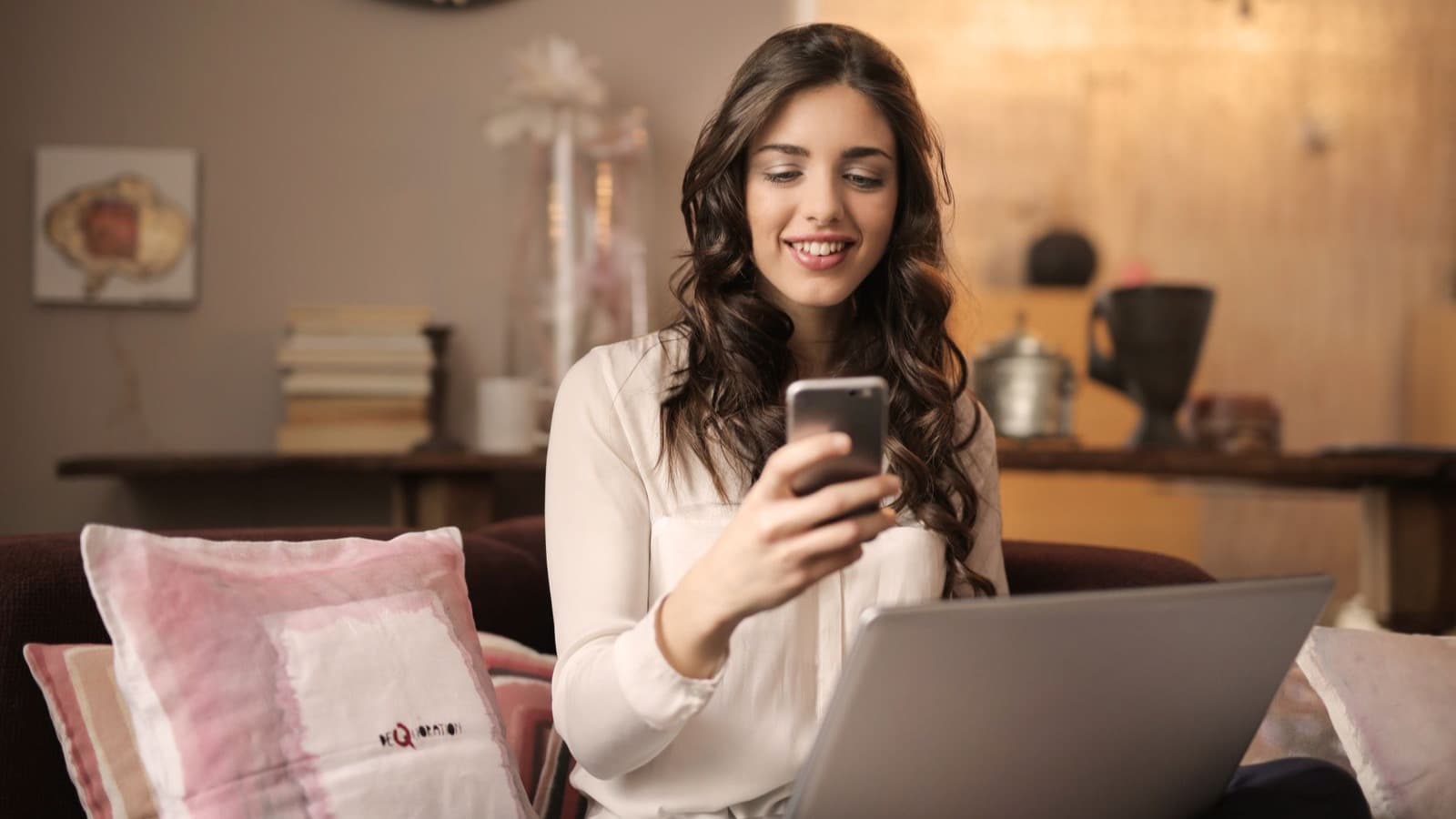 Mulher sorrindo com celular e laptop em casa, sob luz quente