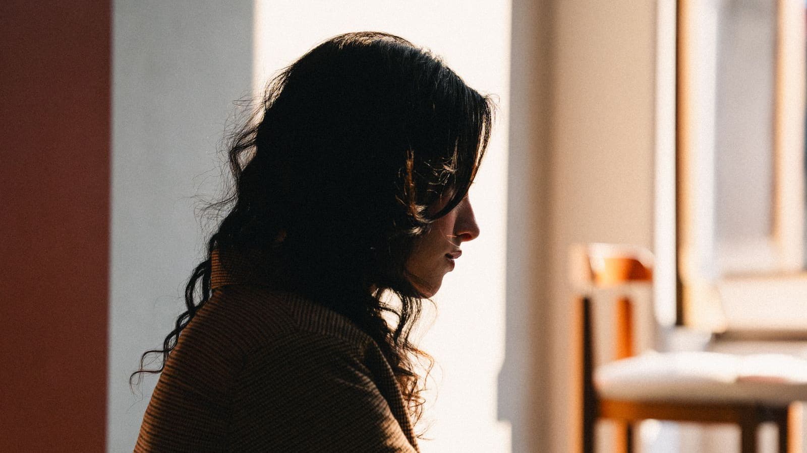 Mulher de perfil junto à janela, iluminada por luz quente, em atitude reflexiva