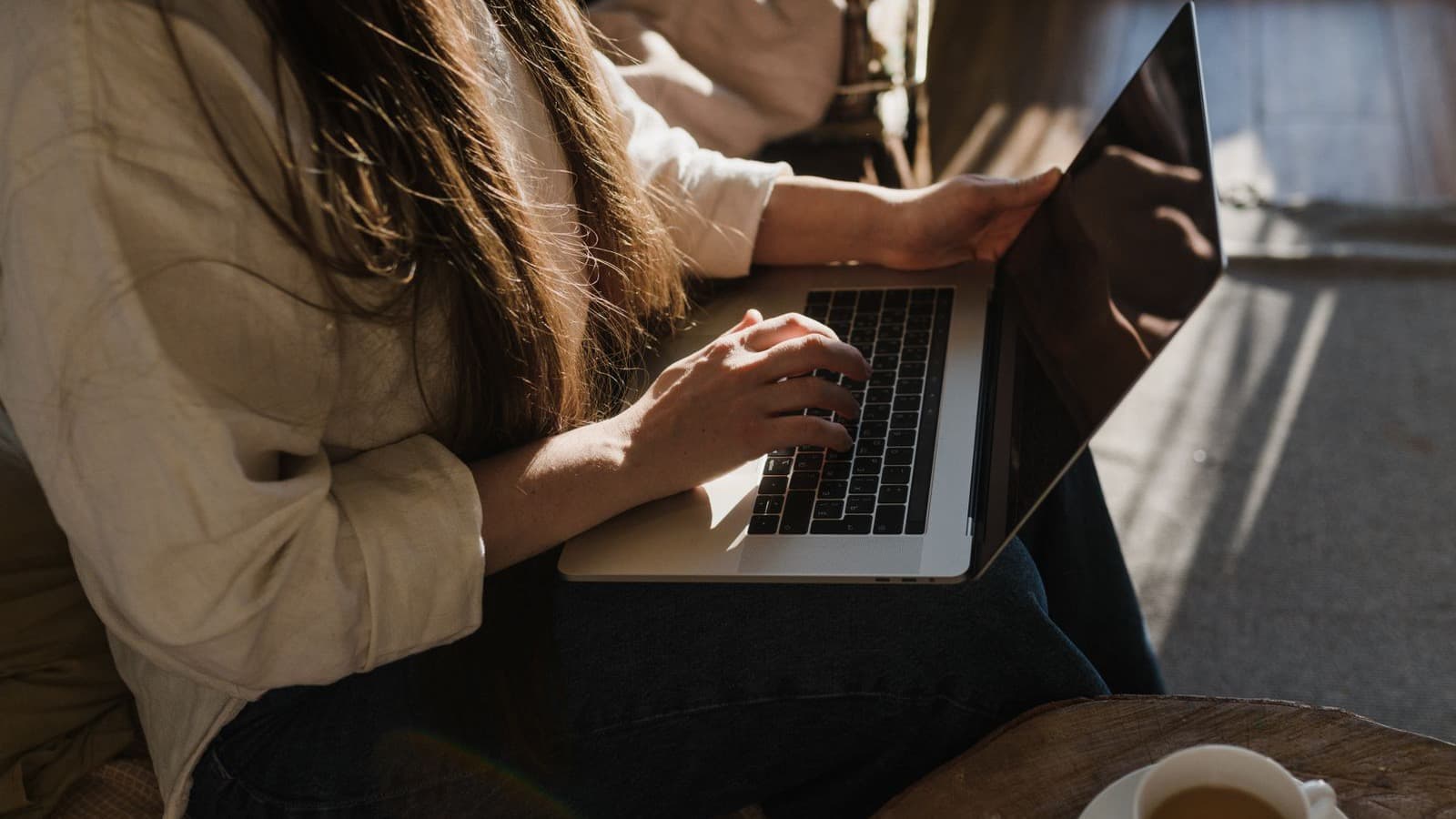Mãos sobre um laptop ao lado de uma xícara de chá, em ambiente aconchegante