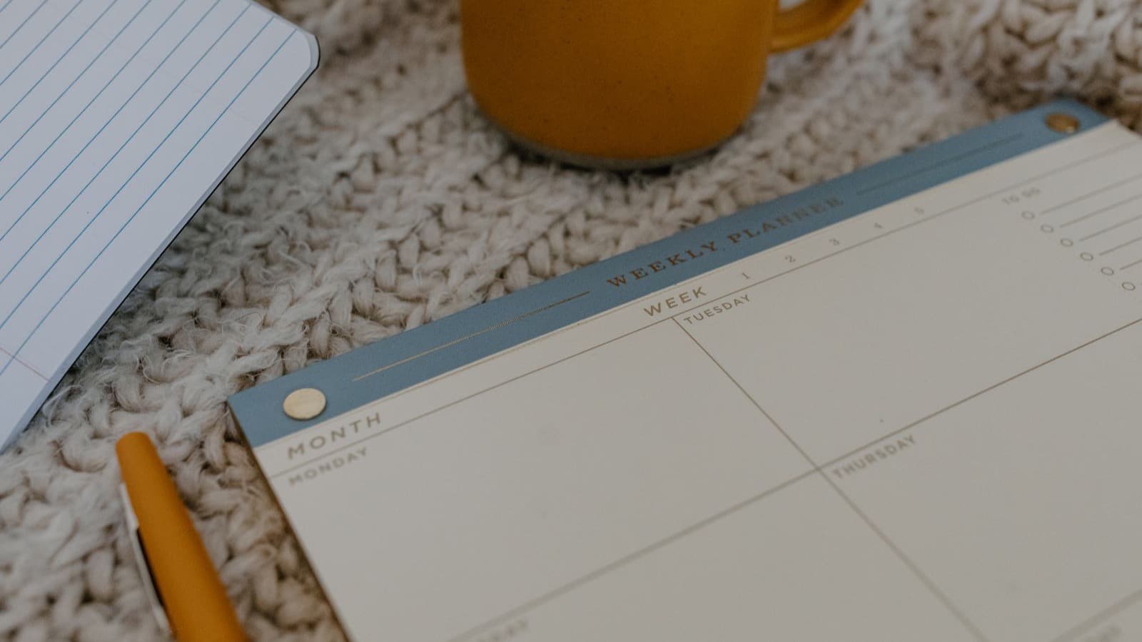 Planner semanal aberto ao lado de uma caneca âmbar e um tricô